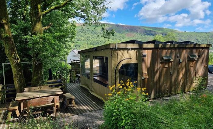 A wooden cabin with a picnic table surrounded by trees and a backdrop of a mountain and blue sky